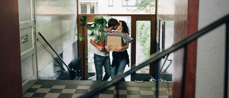 Ett ungt par på väg upp i ett trapphus kysser varandra med händerna fulla av flyttlådor och blommor.
