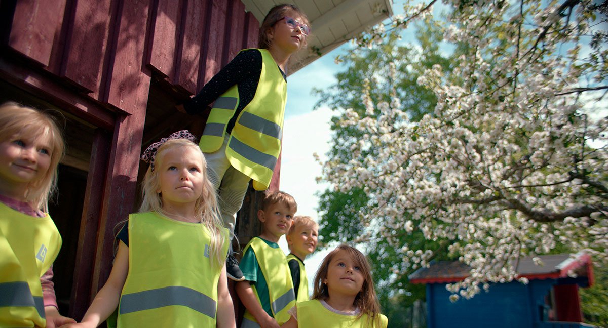 Ett gäng förskolebarn iförda gula reflexvästar står vid en röd byggnad med blommande äppelträd bredvid.