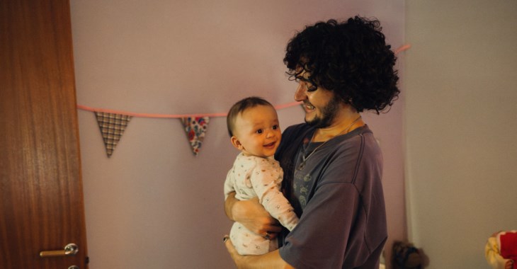 Foto på en pappa med bebis i famnen som har ett samtal med  en mamma som står vid vasken i köket.