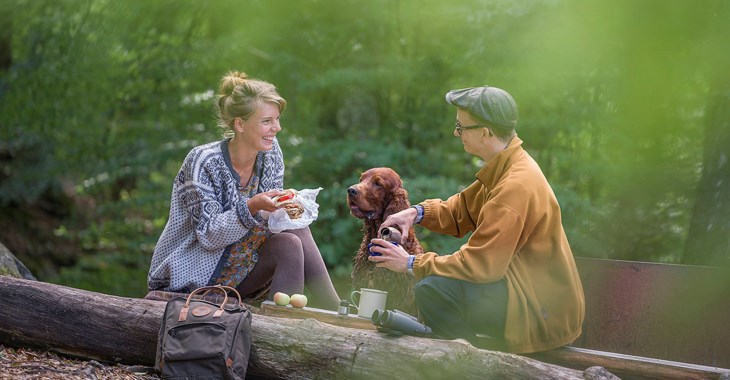 En tjej och kille fikar i skogen tillsammans med sin hund.