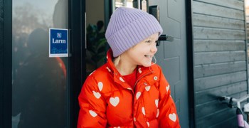 Glad, liten tjej som står utanför en ytterdörr med en larmdekal från trygghetstjänsten Alf.