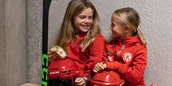 Två glada flickor i röda hockeykläder med var sin hjälm i knät sitter och tittar på varandra.