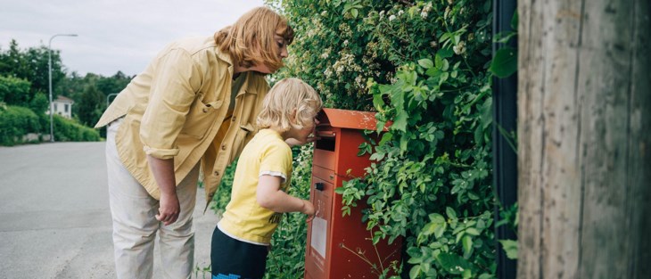 En mamma med sitt lilla barn står och kikar ner i en röd brevlåda.