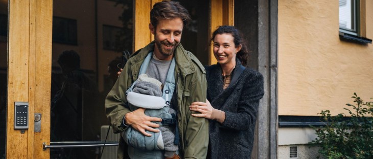 En mamma och en pappa med en bebis i en bärsele på magen, har precis gått ut genom en lägenhetsport.