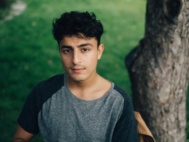 En ung man står vid ett träd i en grönskande park och tittar in i kameran.