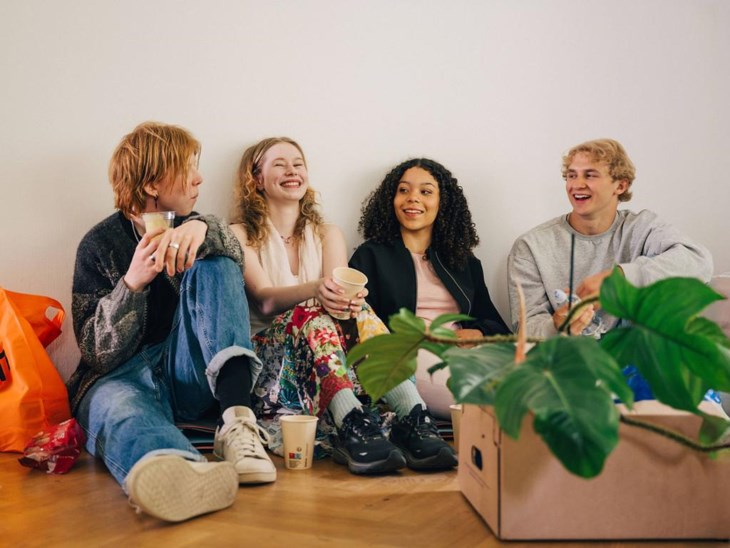Ett kompisgäng sitter på golvet och skrattar i en ny lägenhet, med flyttlådor runt sig.