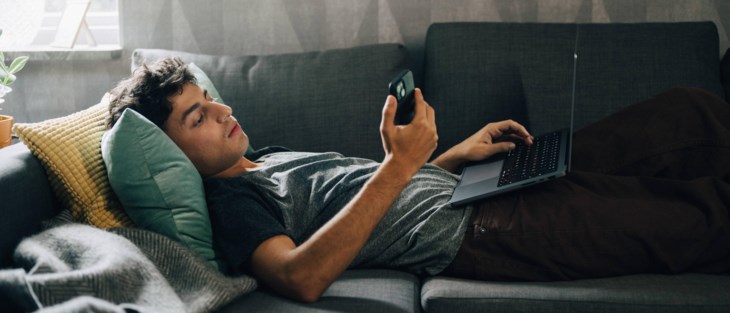 Ung man ligger i soffa med telefon och dator.