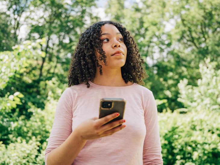 En ung kvinna i rosa tröja blickar upp från sin mobil som hon håller i handen.
