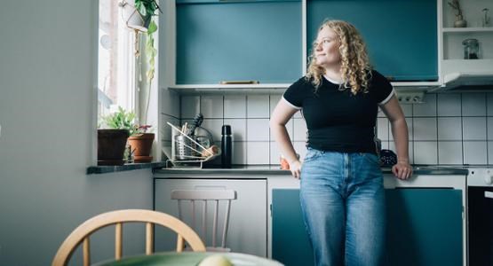 En kvinna står och lutar sig mot köksbänken och blickar ut genom fönstret.