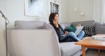 En kvinna sitter i soffan och pratar i mobilen med sin laptop i knät.