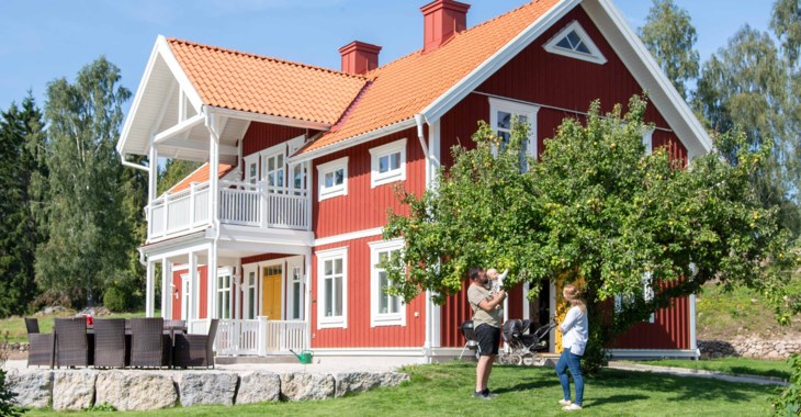 Ett rött trähus med vita knutar, en grön trädgård och två personer vid ett äppelträd.