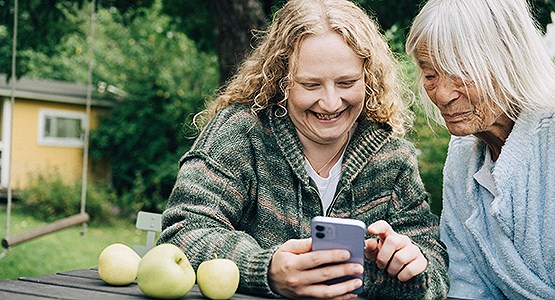 Kvinna sitter med sin farmor och kollar i telefonen. De sitter utomhus med grönska och ett hus i bakgrunden.