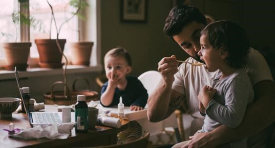 En pappa sitter vid köksbordet i dunkelt ljus och ger ett av sina två barn medicin.