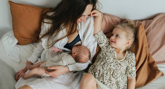 En nyfödd bebis ammar hos sin mamma, storasyster sitter bredvid.