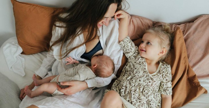 En nyfödd bebis ligger i sin mamma famn och ammar, storasyster sitter bredvid.