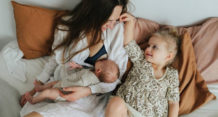 En mamma sitter i sängen och ammar sin bebis, bebisens storasyster sitter bredvid och myser.