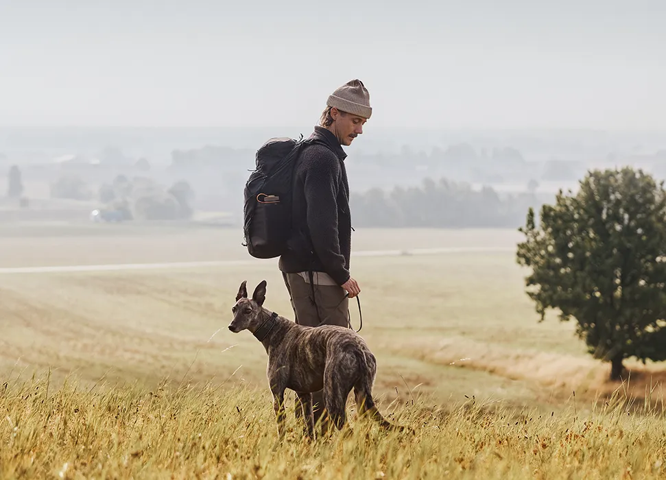 Person med ryggsäck står på en äng och håller i en hund med landskap i bakgrunden.