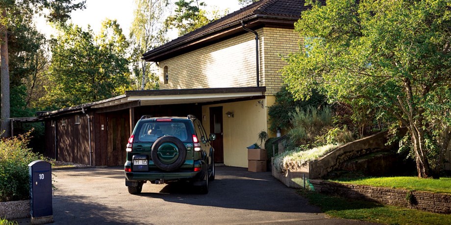 En mörk SUV står parkerad på en asfalterad uppfart framför ett gult tvåvåningshus med carport, omgiven av gröna träd och buskar i ett villakvarter.