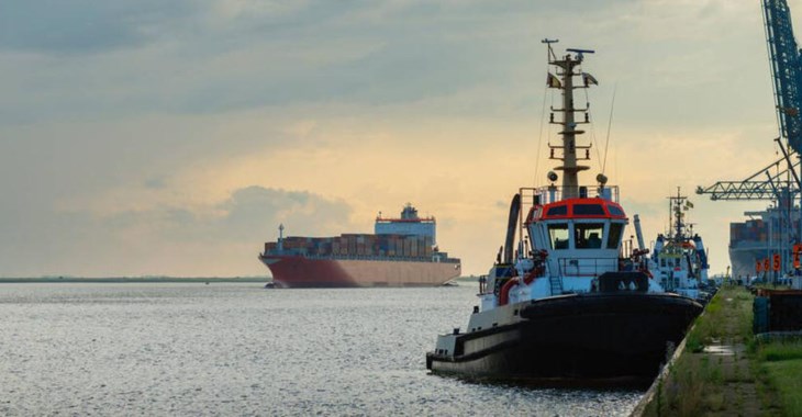 Lotsbåt ligger i hamn och ett stort fraktfartyg är på väg till havs i skymningen.