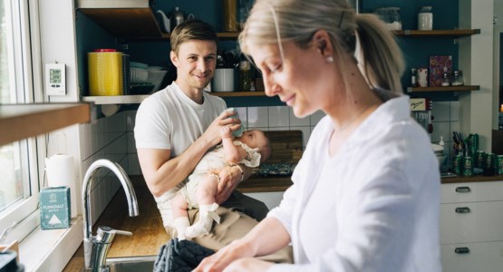 En barnfamilj står i ett kök. Mamman diskar och pappa matar bebis.
