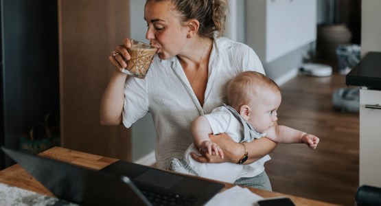 Mamma jobbar vid laptop med bebis i famnen.