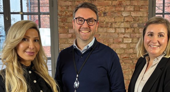 Tre personer står framför en tegelvägg och tittar in i kameran.