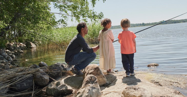 En pappa med två barn står på en berghäll vid vattnet och fiskar en solig sommardag.