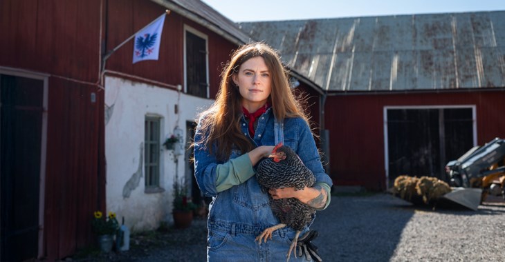 Kvinna framför ladugård som håller en höna.
