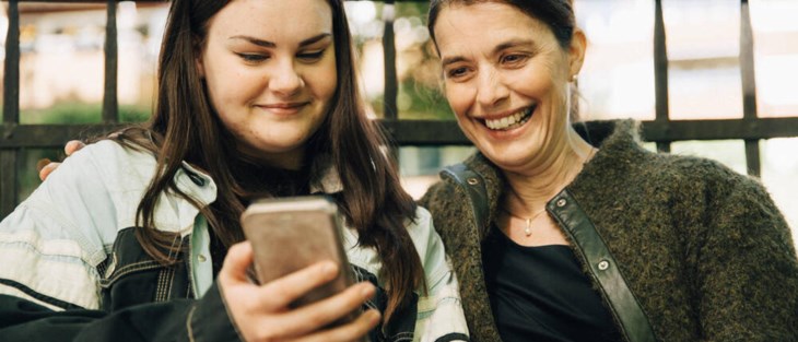 Mamma och tonårsdotter tittar på  något i mobiltelefon.
