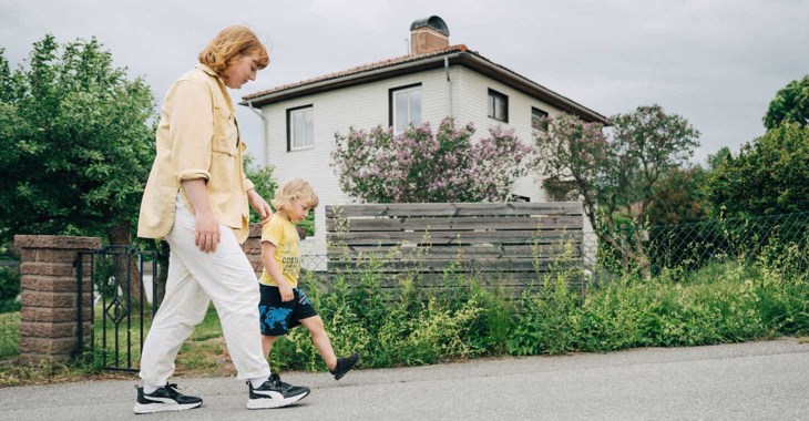 Mamma och son promenerar, med sitt hus i bakgrunden.