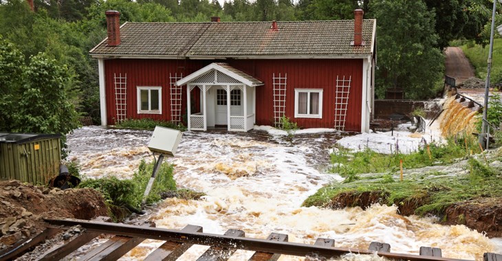 Röd stuga med kraftig översvämning utanför. Ett järnvägsspår syns i förgrunden
