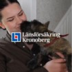 Person i brun skjorta håller en liten hund med röd sele.