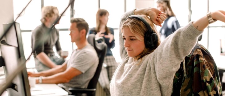 En kvinna med hörlurar sträcker på sig framför datorn.