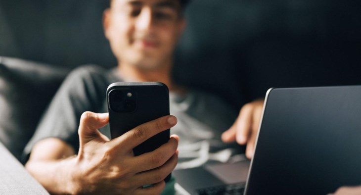En man sitter i soffan framför en dator och tittar på sin mobil.