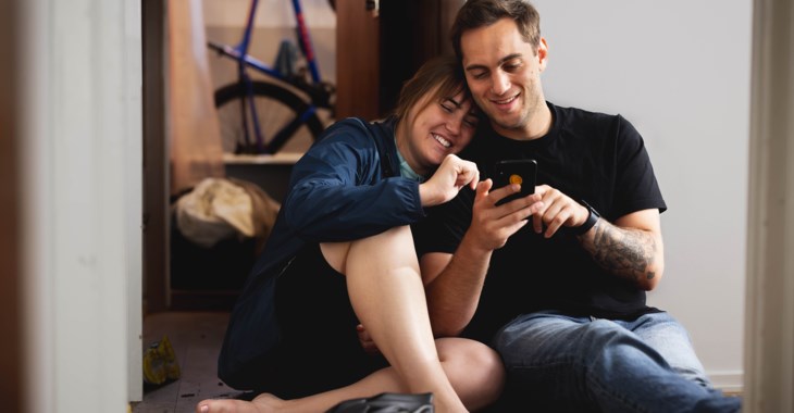 Ungt par sitter på golvet och tittar på en mobiltelefon.