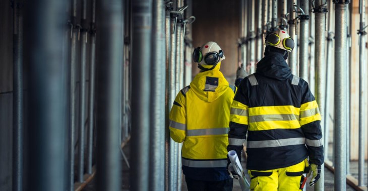 Två arbetsklädda män går på byggarbetsplats med ryggen mot kameran.