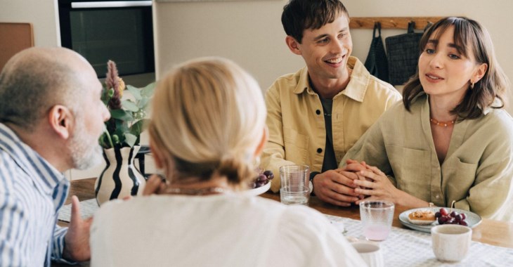En familj med äldre föräldrar och ett vuxet barn med sin aprtner, sitter och samtalar vid ett matbord.