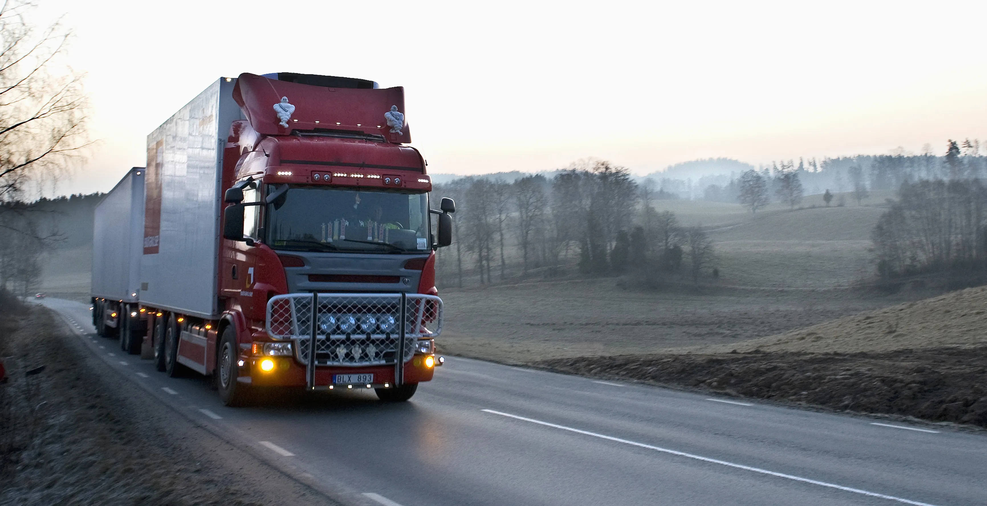 En röd tung lastbil dundrar förbi på en landsväg en disig vårmorgon någonstans i Sverige.