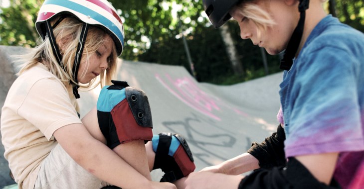 Två barn med hjälmar och skydd sitter vid en skatepark, ett hjälper det andra.