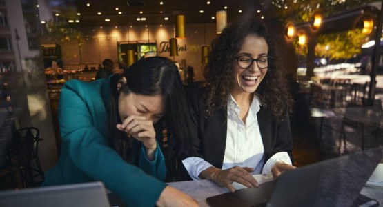 två kvinnor sitter framför en dator på ett café och skrattar