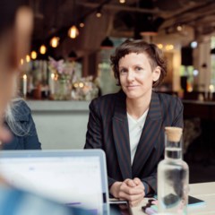 Kvinna i kavaj sitter vid ett bord på en restaurang. Kvinnan tittar in i kameran.