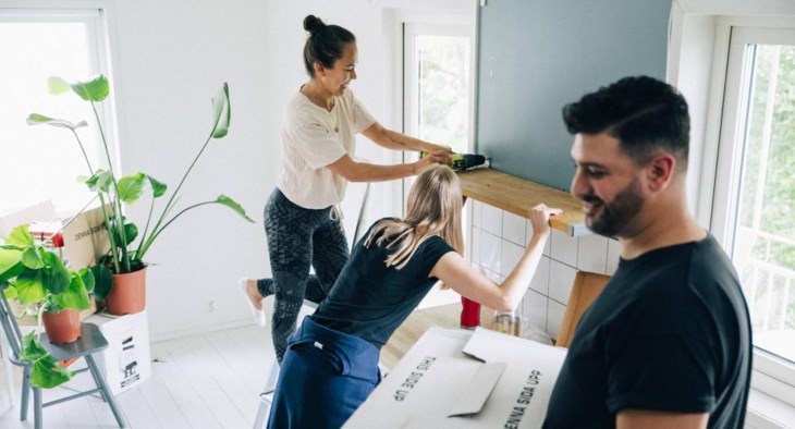 Två kvinnor sätter upp en hylla i sitt nya hem, en kompis hjälper till att bära en flyttlåda.