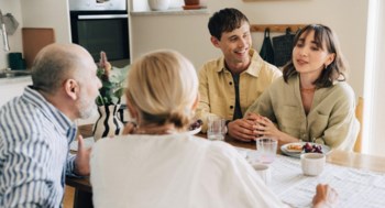 En familj med två äldre föräldrar, ett vuxet barn och dennes sambo sitter vid matbordet och pratar.