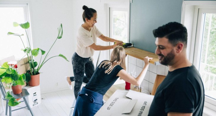 Två kvinnor skruvar upp en hylla i ett kök medan deras kompis bär flyttlådor.