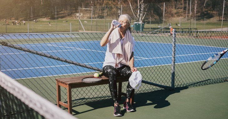 Äldre dam dricker vatten på tennisbana