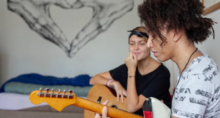 Två syskon i tonåren sitter på en säng och spelar gitarr.