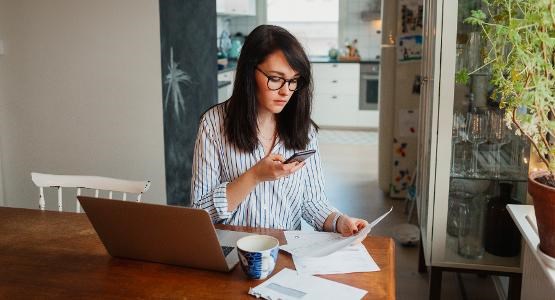 Kvinna sitter vid sin bärbara dator. Hon har en mobiltelefon i handen och papper ligger på skrivbordet. 