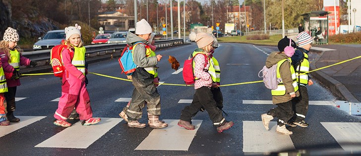 Förskolebarn gåendes i reflexvästar på ett övergångsställe över en trafikerad väg.